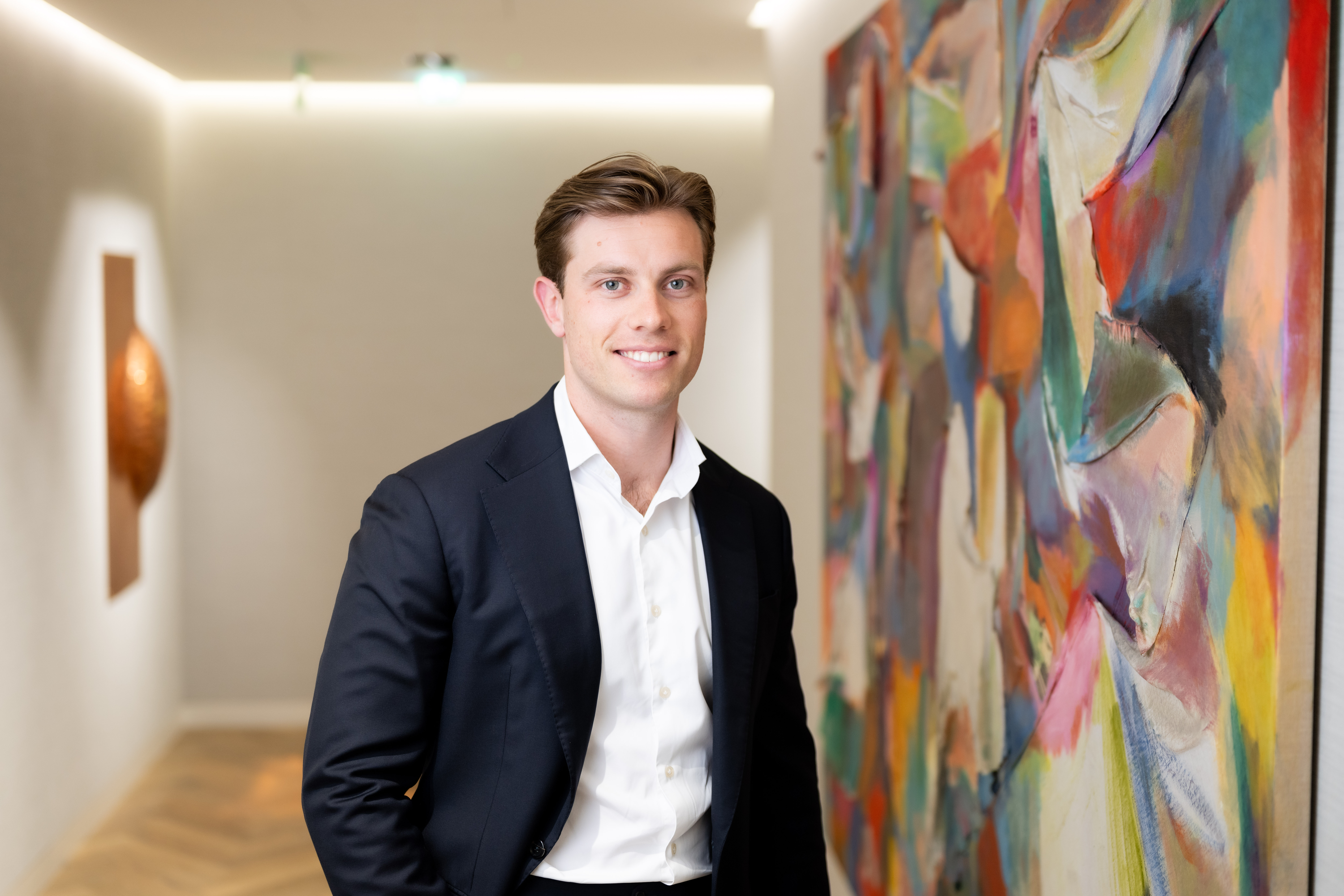 Professional portrait of a person wearing a dark suit jacket and white shirt, smiling beside a colorful abstract painting in a modern office.