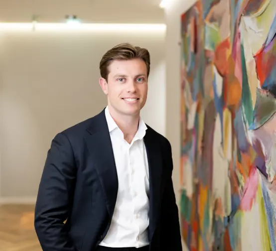 Professional portrait of a person wearing a dark suit jacket and white shirt, smiling beside a colorful abstract painting in a modern office.