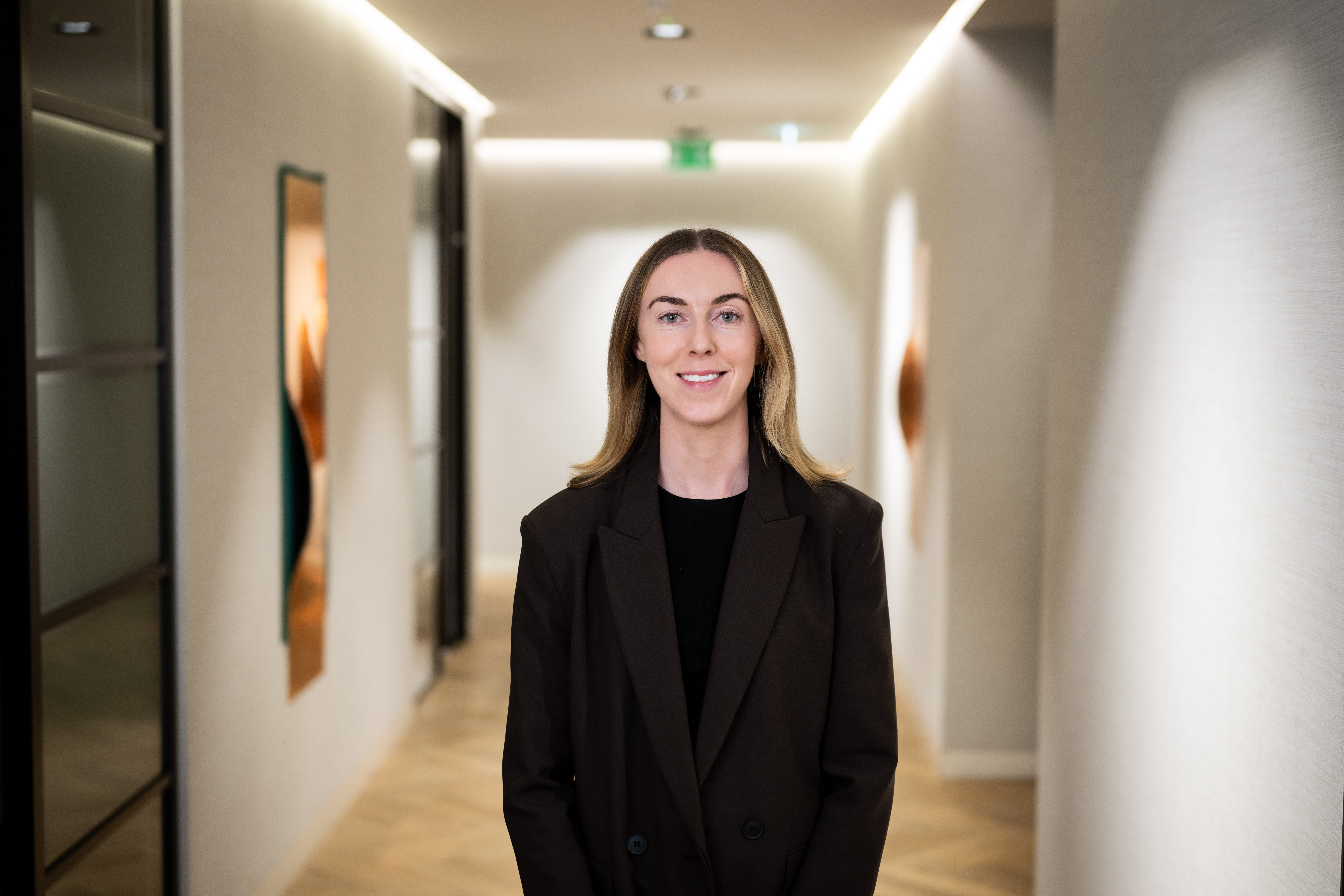 Professional portrait of a person wearing a dark blazer over a black top, smiling at the camera in a modern office hallway.