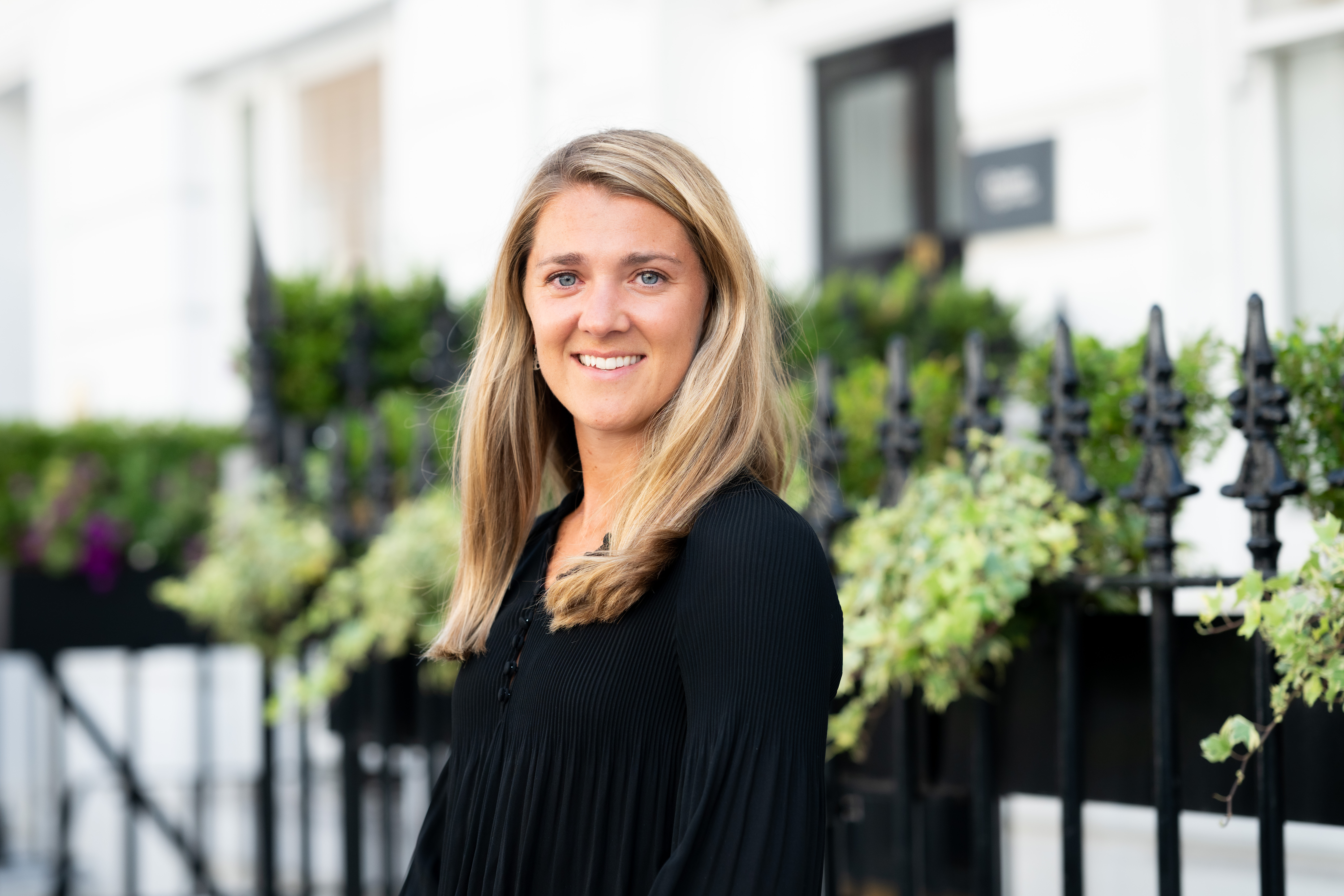 A professionally photographed portrait of a person with shoulder-length blonde hair wearing a black top, smiling at the camera. They are standing outdoors beside a black wrought-iron fence with green foliage, with a softly blurred white building in the background.