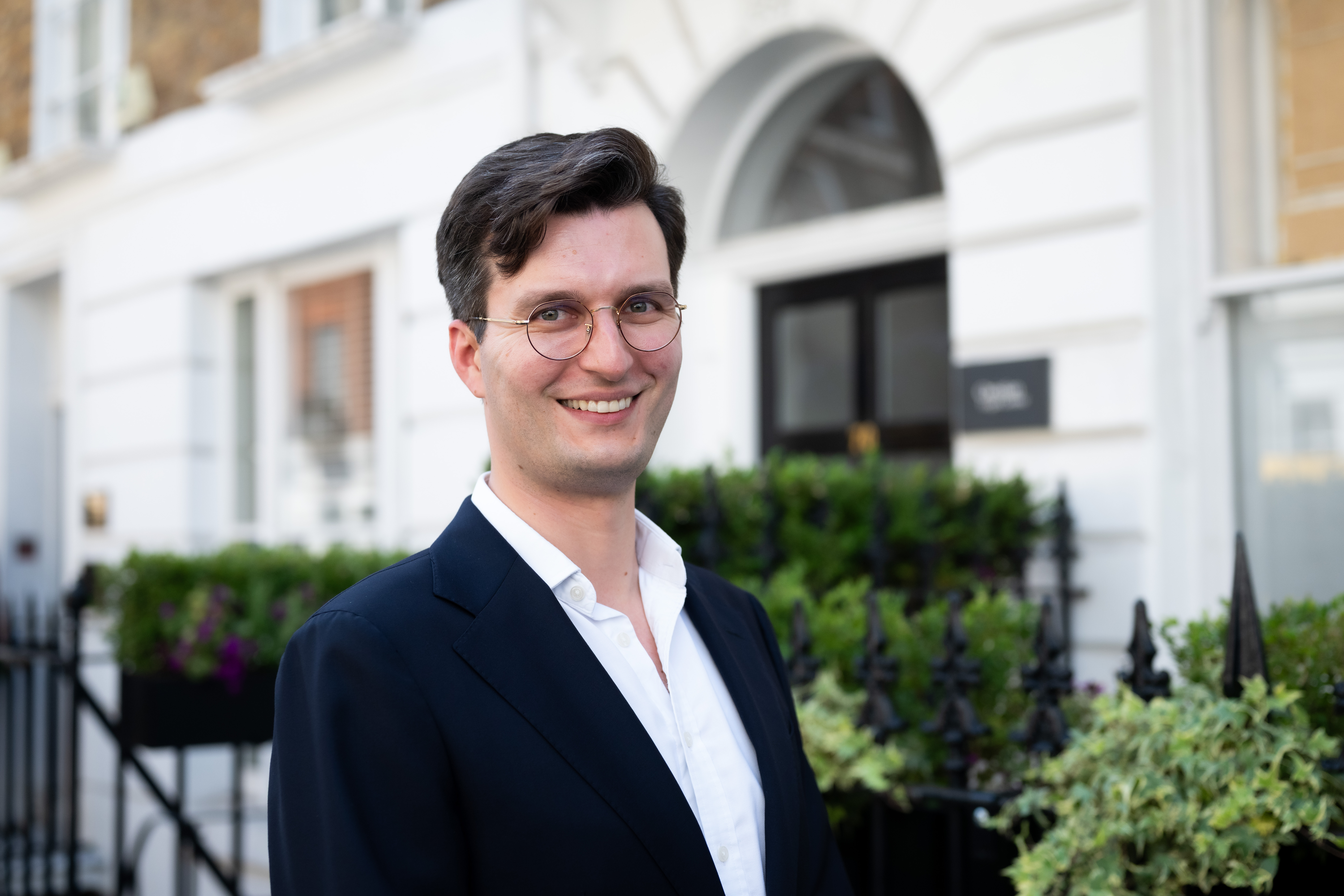 A professional headshot of a smiling man with dark hair and round glasses, dressed in a navy blazer and white open-collar shirt, standing outside a white Georgian-style building with an arched doorway, black railings, and landscaped greenery.