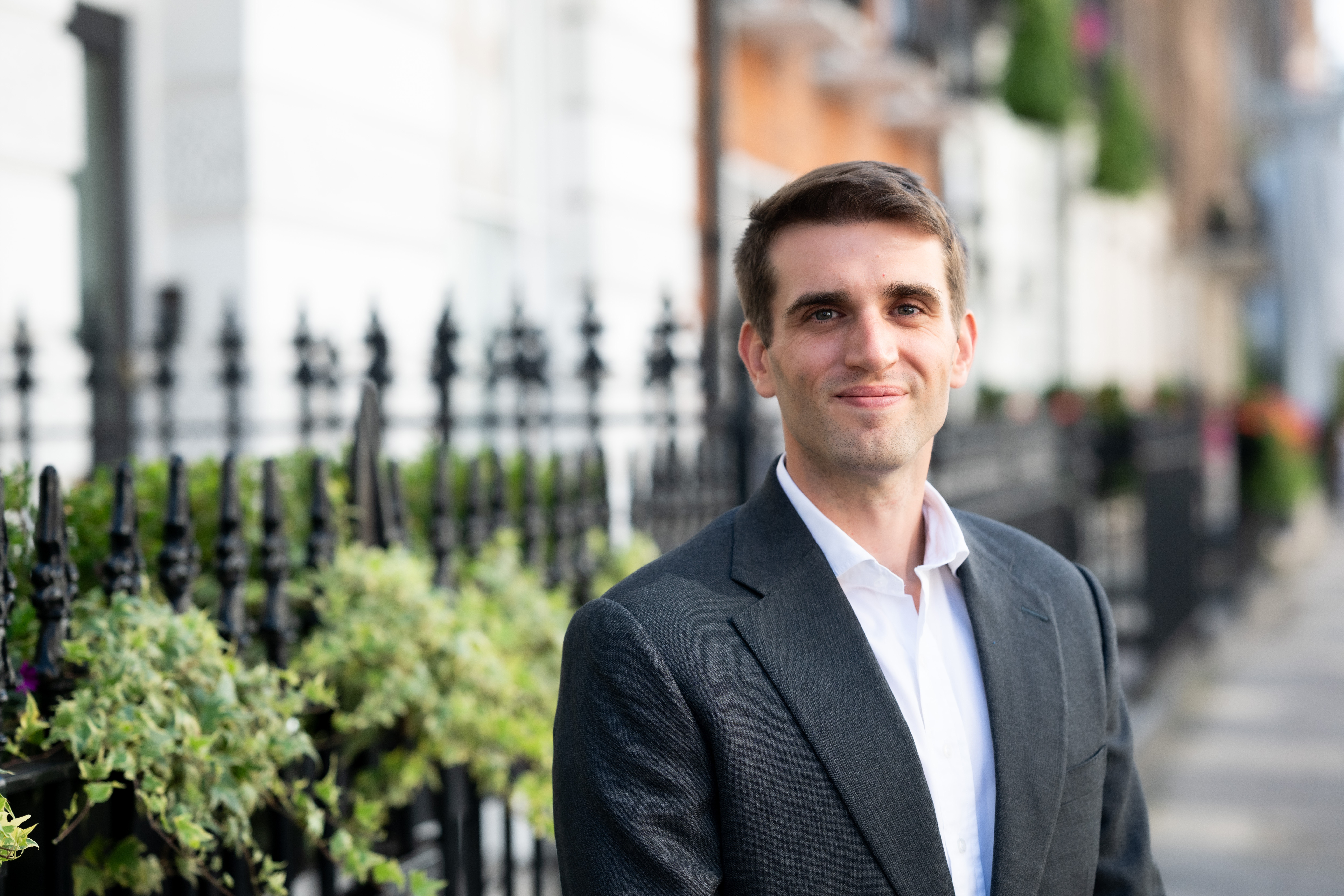 Professional outdoor headshot of a person in business attire smiling at the camera, with elegant townhouses and greenery softly blurred in the background.