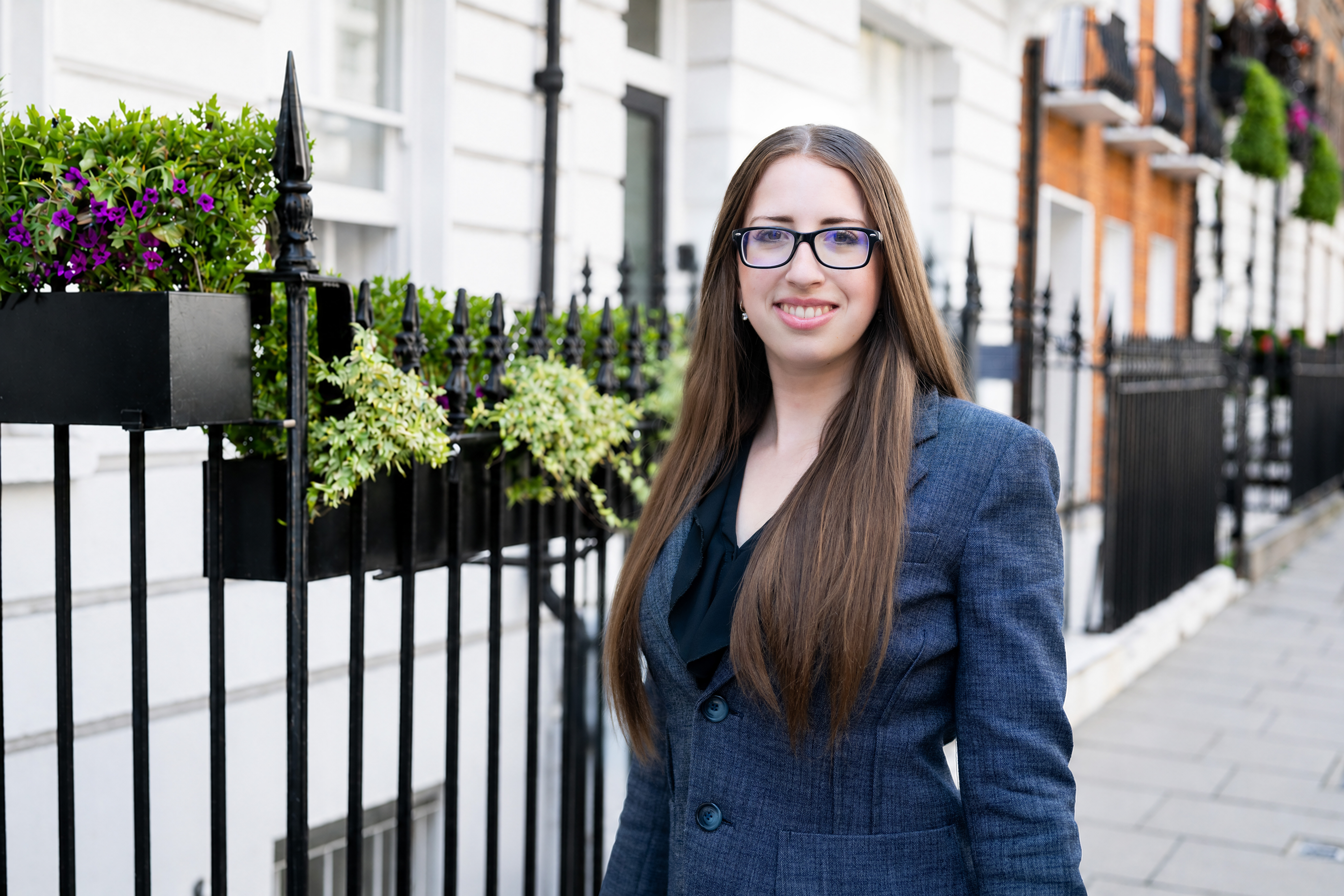 Professional headshot of a woman standing on a London street, wearing a navy blazer and glasses, smiling at the camera.