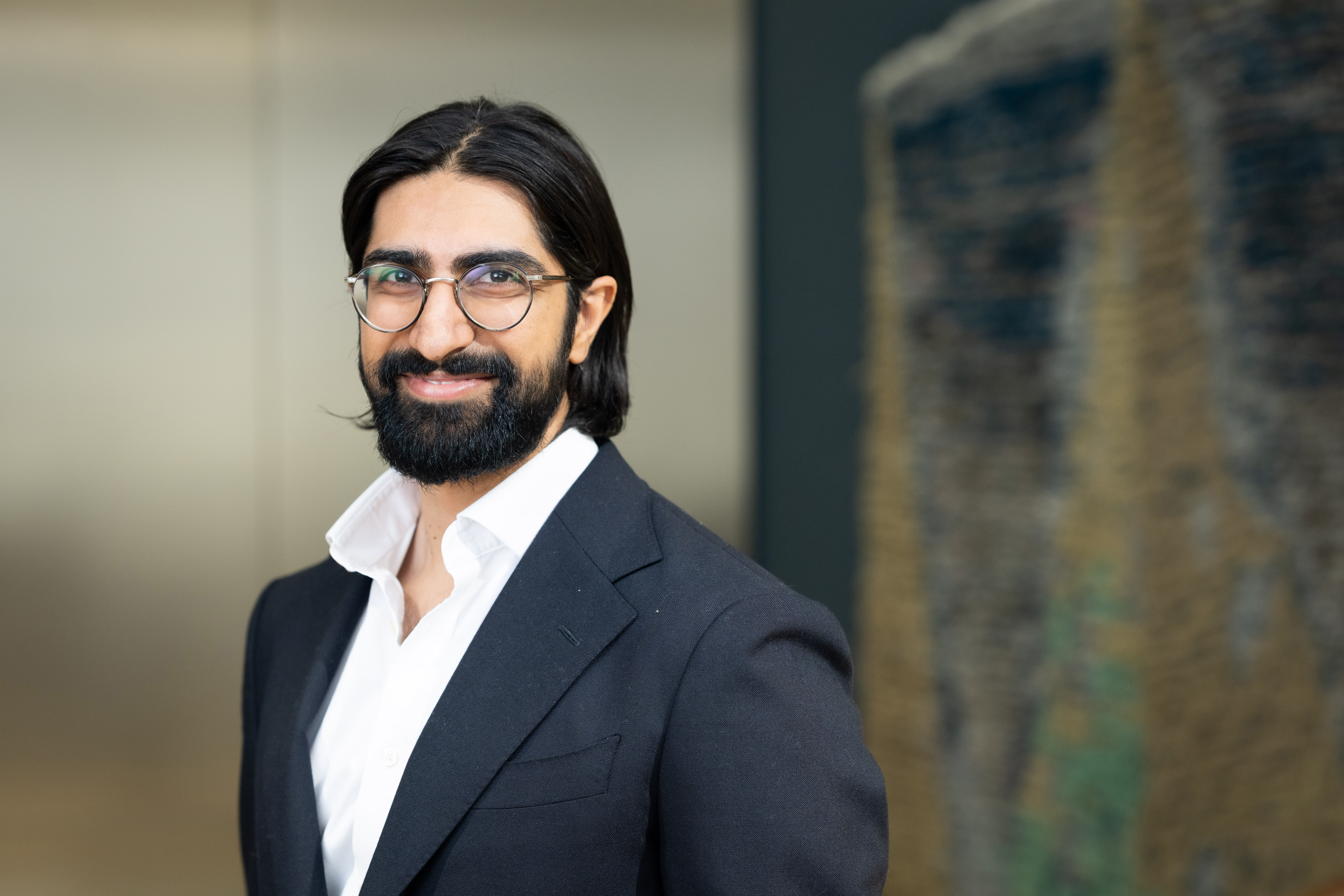 Professional headshot of a man with long dark hair, glasses, and a beard, wearing a dark blazer and white shirt, smiling at the camera.