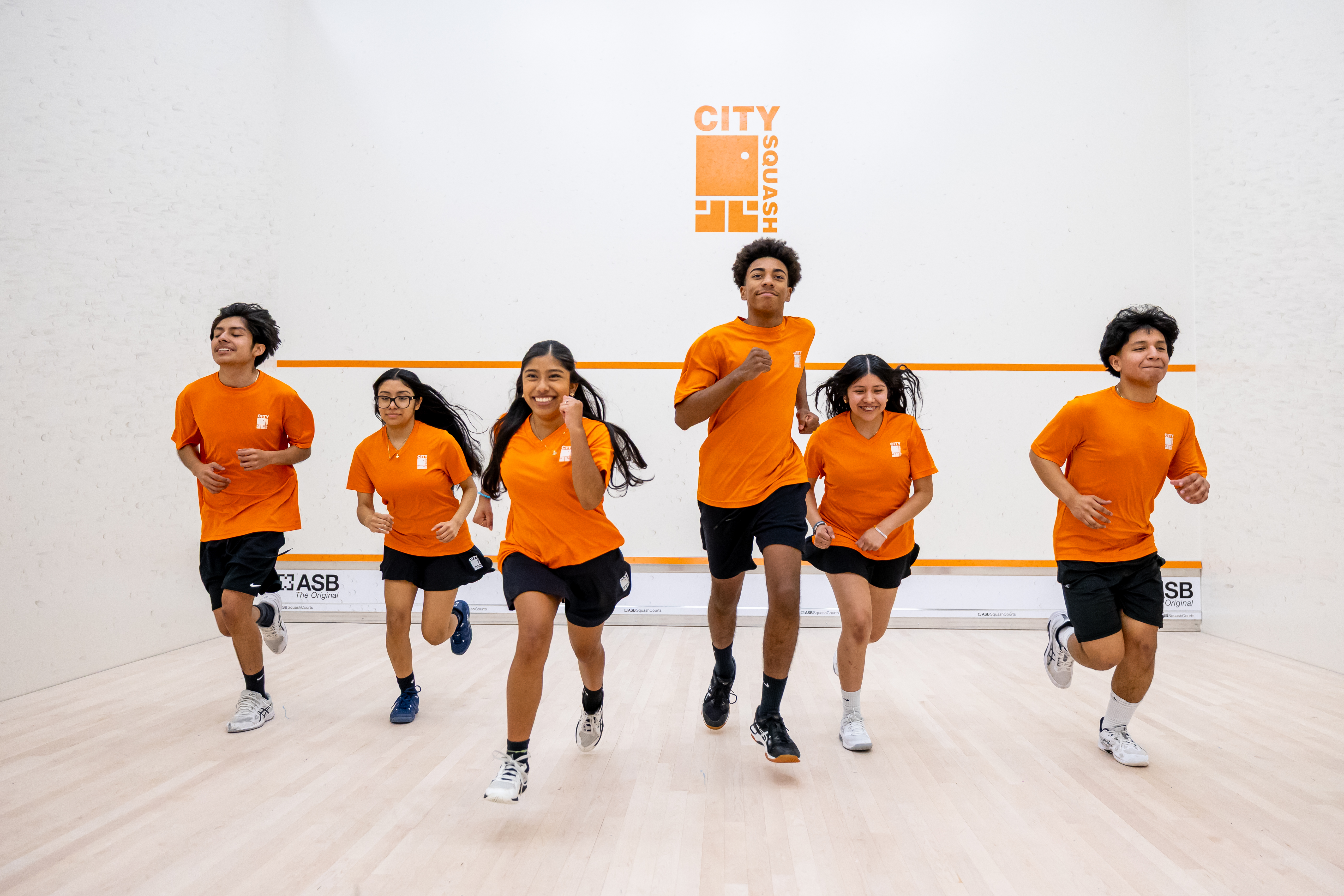 Six young people in orange City Squash kit running towards the camera on a squash court.