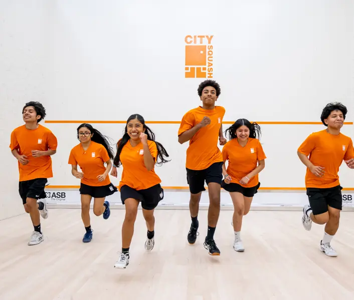Six young people in orange City Squash kit running towards the camera on a squash court.