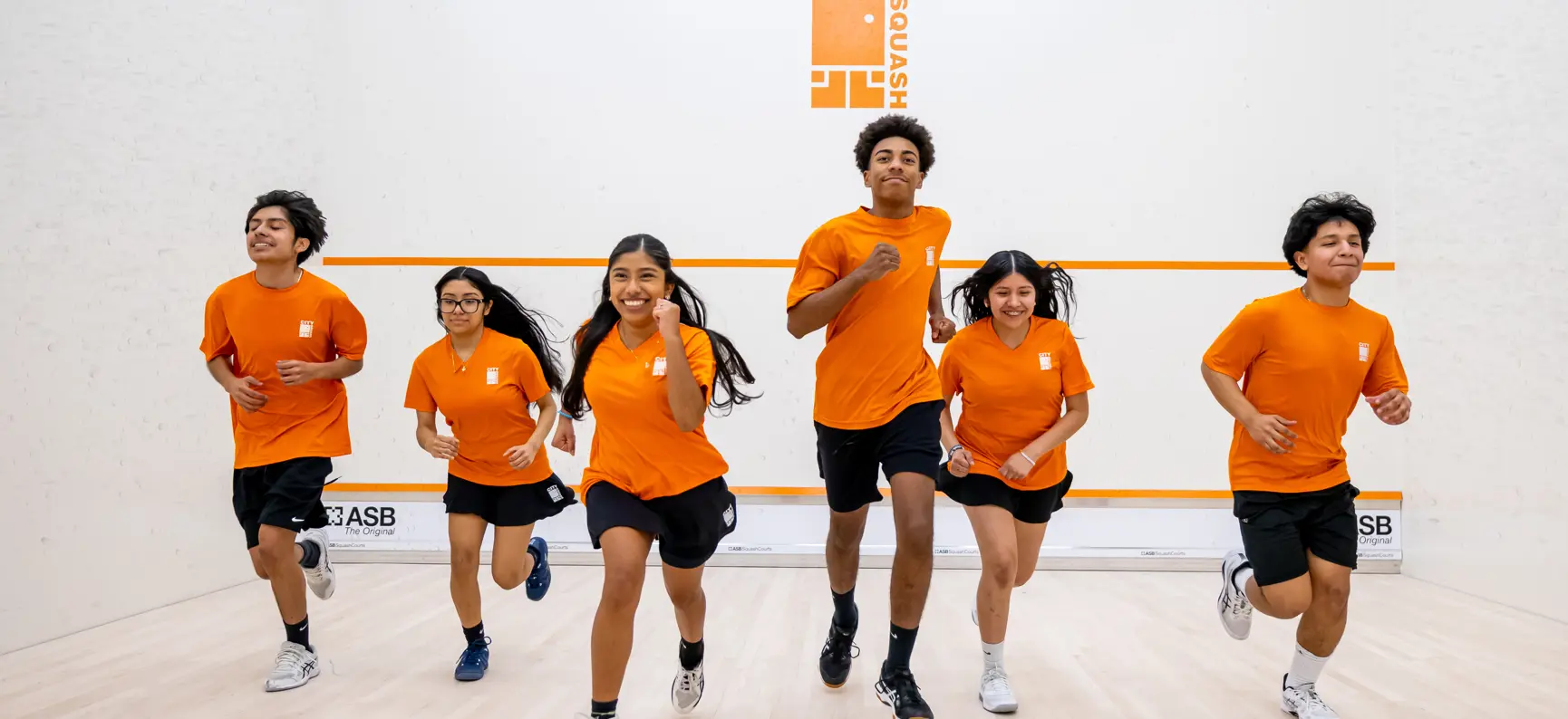 Six young people in orange City Squash kit running towards the camera on a squash court.