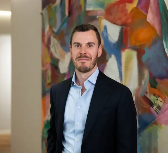 Professional headshot of a man in business attire standing in front of a colorful abstract artwork in a modern office setting.