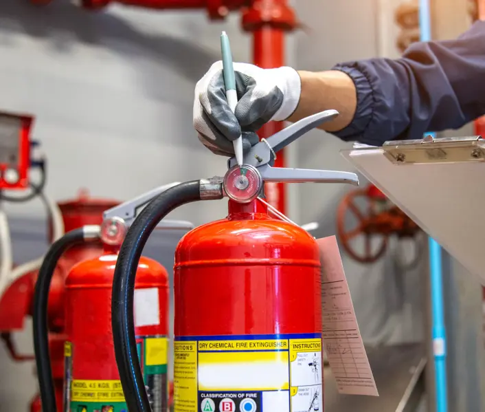 A fire safety technician inspecting a red fire extinguisher in a plant room, holding a clipboard