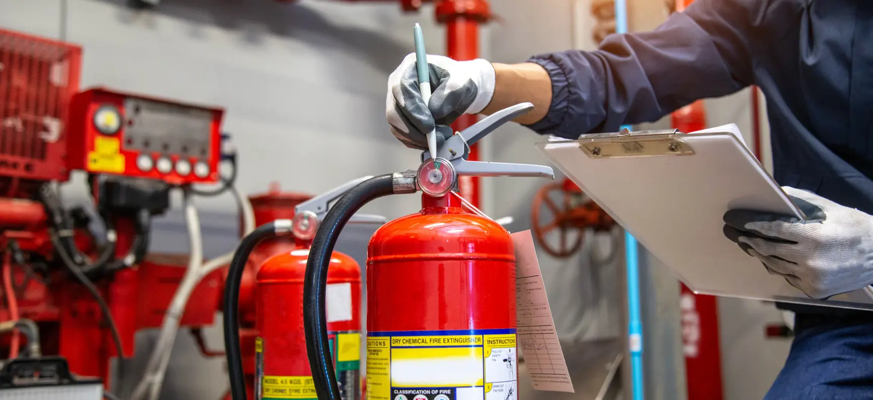 A fire safety technician inspecting a red fire extinguisher in a plant room, holding a clipboard