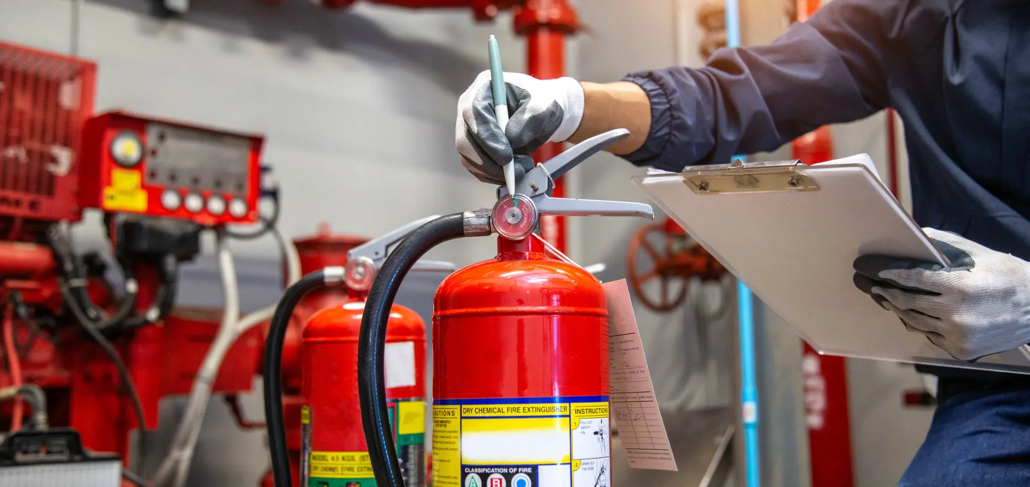 A fire safety technician inspecting a red fire extinguisher in a plant room, holding a clipboard