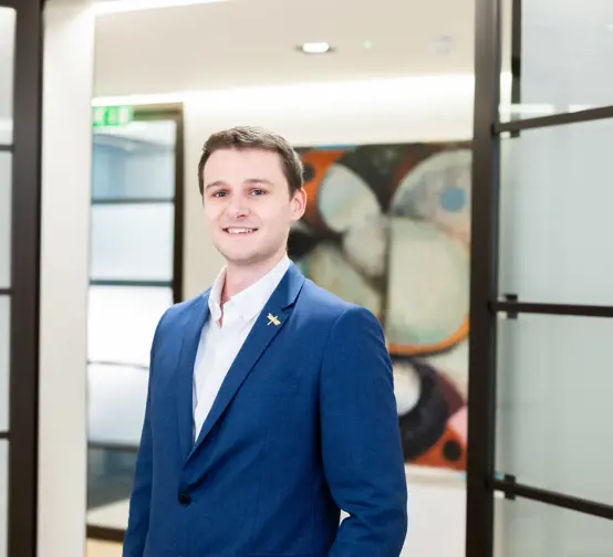 Professional headshot of Connor Rayson standing in the Inflexion office, wearing a blue suit jacket and white shirt.