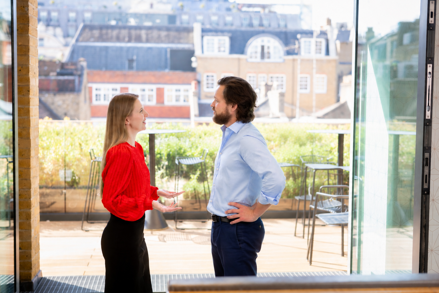 Woman in a red top and man in a light blue shirt talking on a sunlit terrace