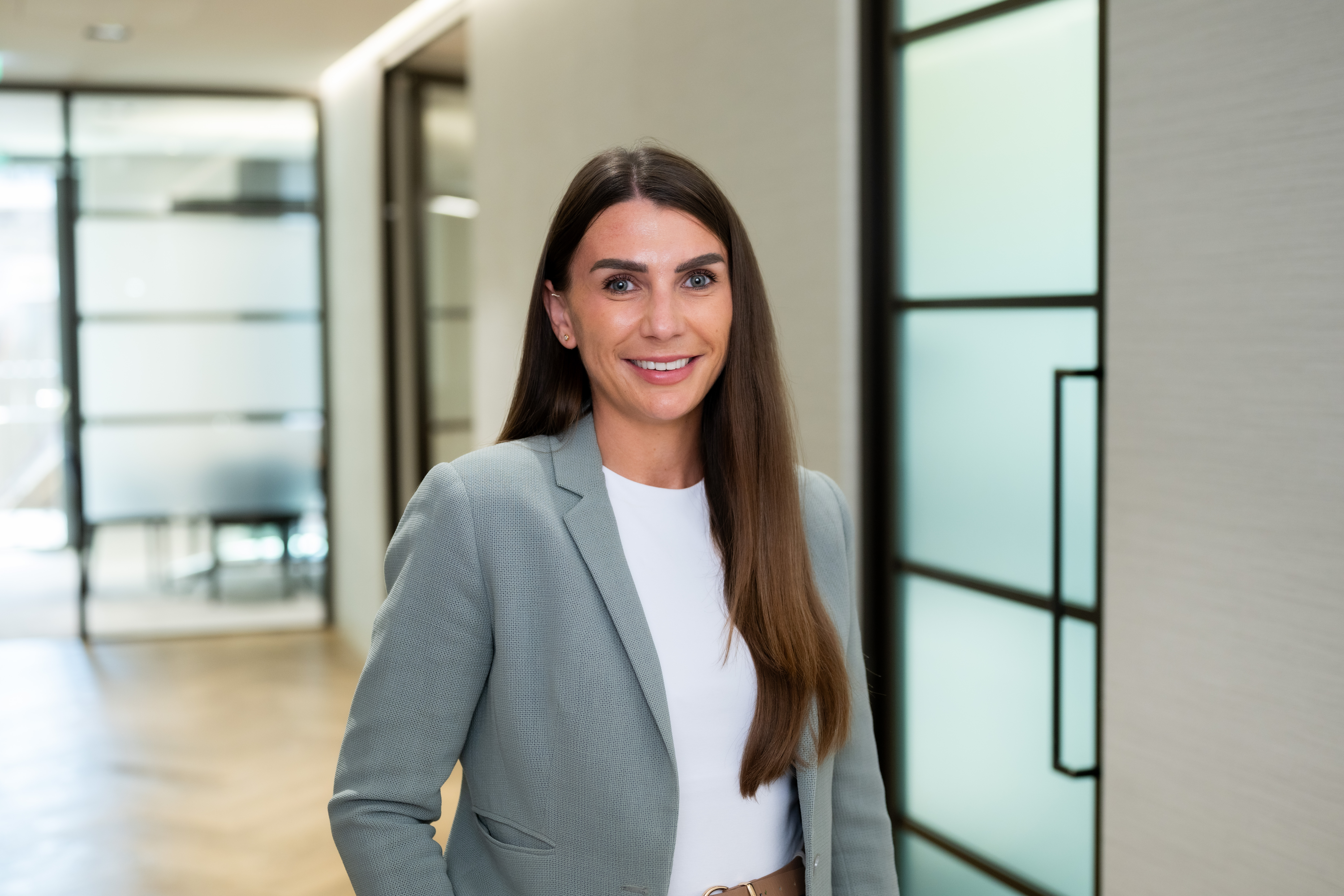 Professional portrait of a team member smiling in a modern office environment, wearing a light grey blazer and white top.
